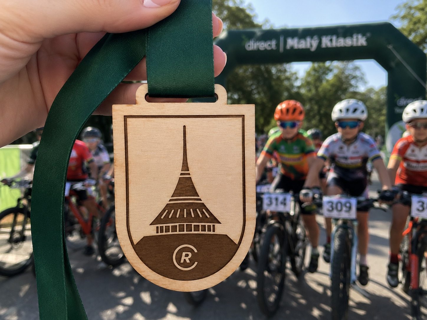 Cyklisté se startují na závodě, v popředí dřevěná medaile HOORAY